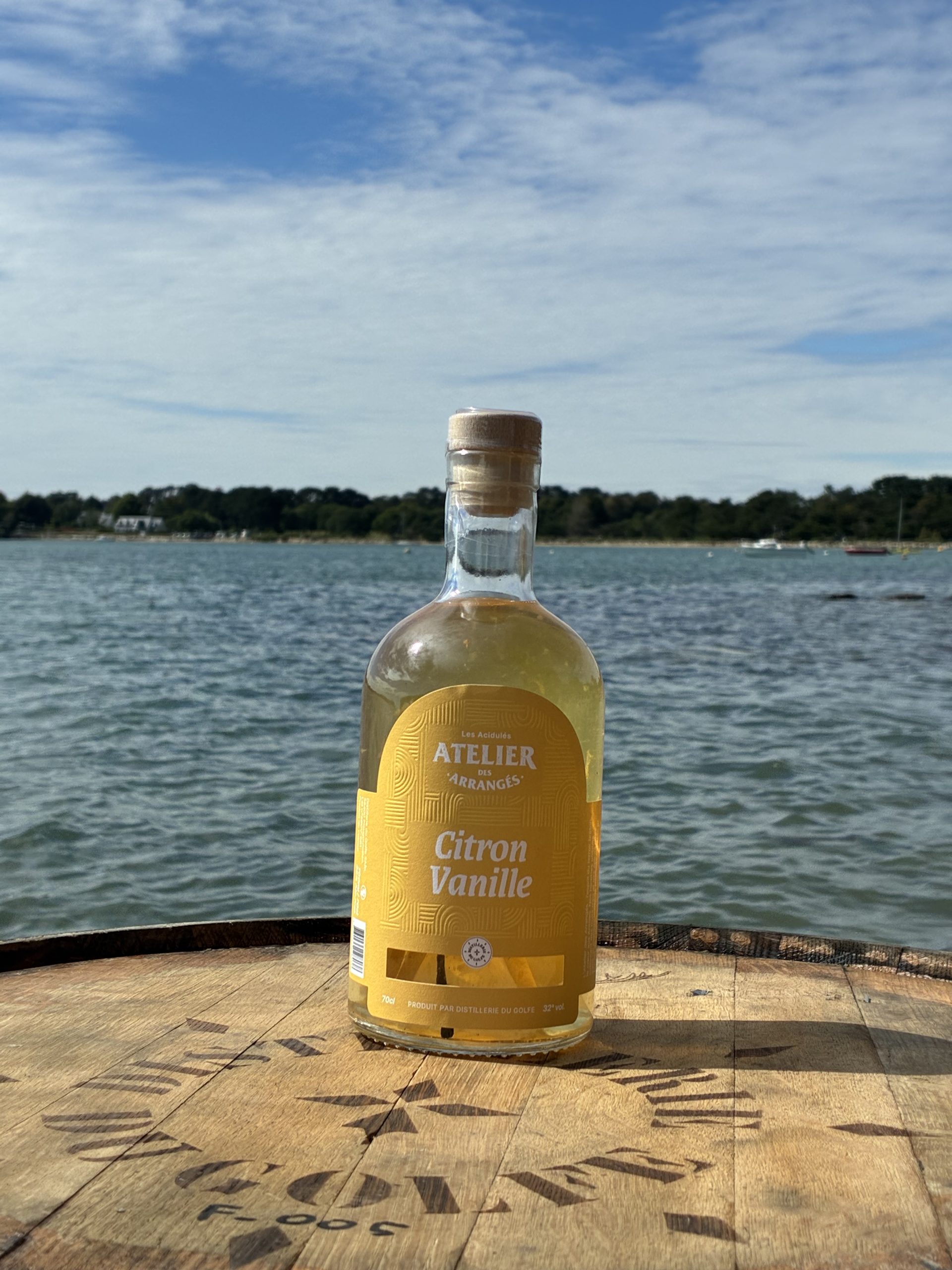 Bottle of Citron Vanille liqueur on a weathered wooden table by a lake, with a blue sky and distant shoreline in the background.