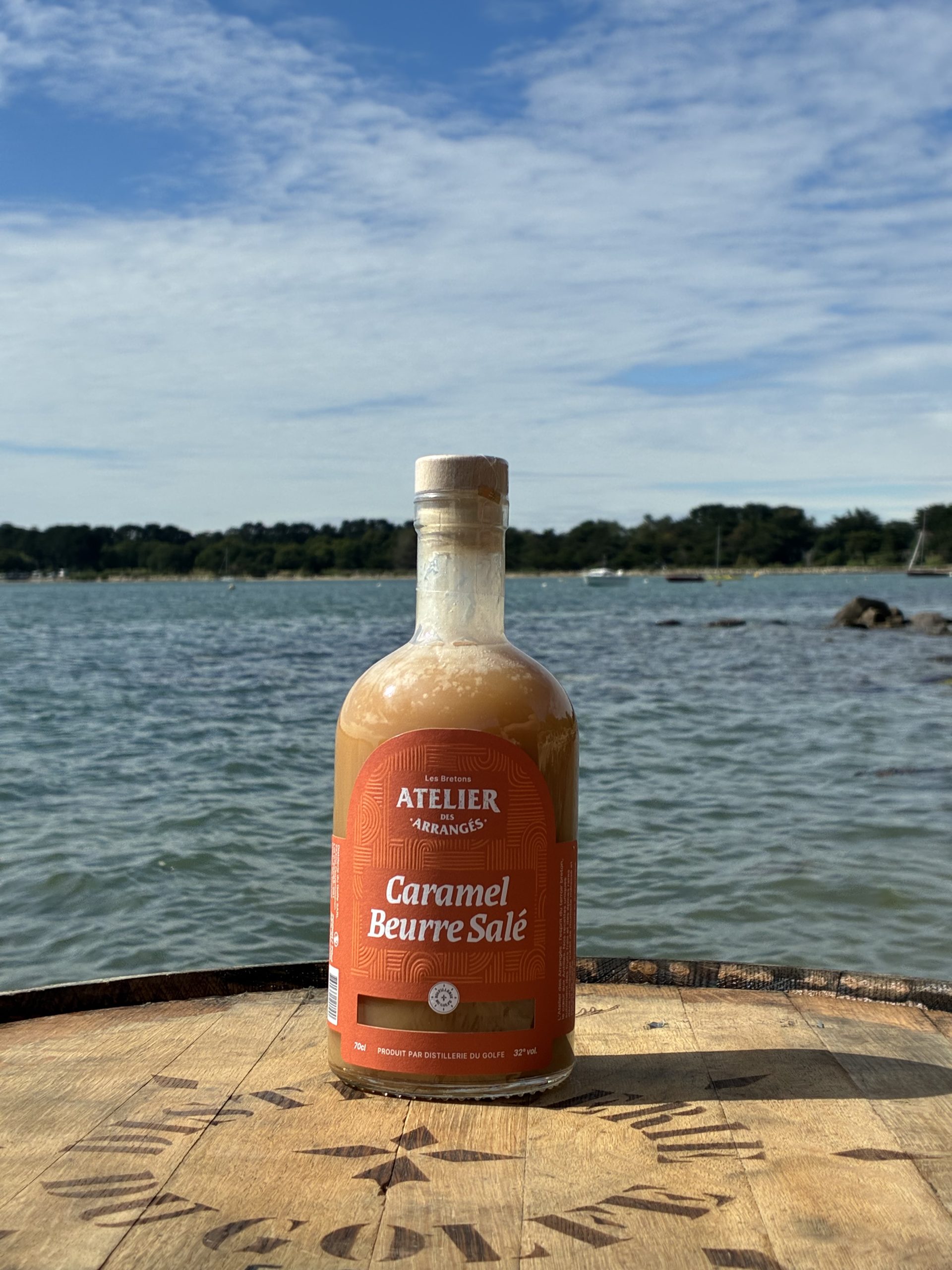 Bottle of caramel beurre salé liqueur labeled Atelier des Arrangés on a wooden dock with water and shoreline in the background.