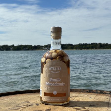 Bottle of Vanille Macadamia liqueur on a wooden table by a lake with a blue sky in the background, label visible.