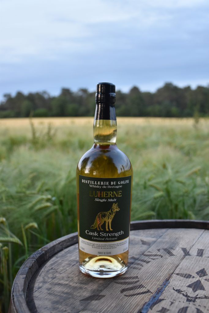 Whiskey bottle on a wooden barrel outdoors in a grassy field with trees in the distance.